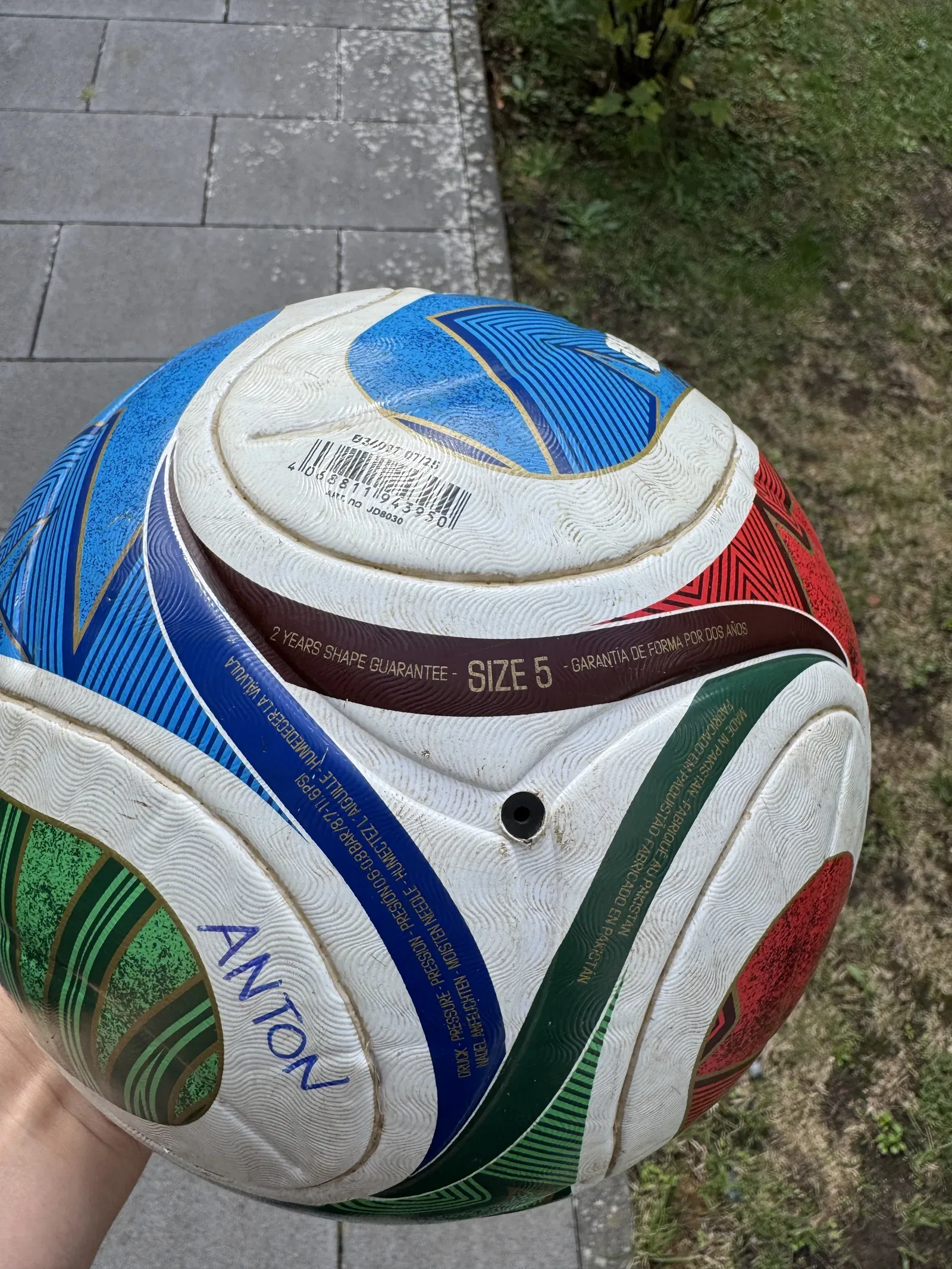 Nahaufnahme eines weißen Fußballs mit grünen, roten und blauen Bahnen. Lesbar sind ein Strichcode, das braune Band mit 2 YEARS SHAPE GUARANTEE und SIZE 5 sowie das blaue Band mit DRUCK PRESSURE PRESSION 0,6 bis 0,8 BAR, 8.7 bis 11.6 PSI und NADEL ANFEUCHTEN. Rechts daneben das schwarze Ventil