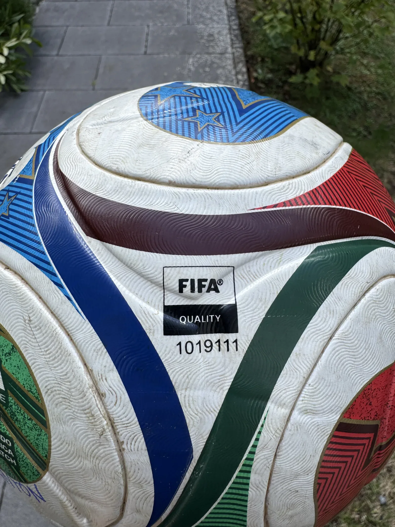 Nahaufnahme der Hülle eines Fußballs über Pflastersteinen. Die weißen Felder sind flächig vergraut und zerkratzt, die goldenen Konturlinien der farbigen Bahnen sind an den Kanten abgerieben. Mittig das Zeichen FIFA QUALITY mit der Nummer 1019111