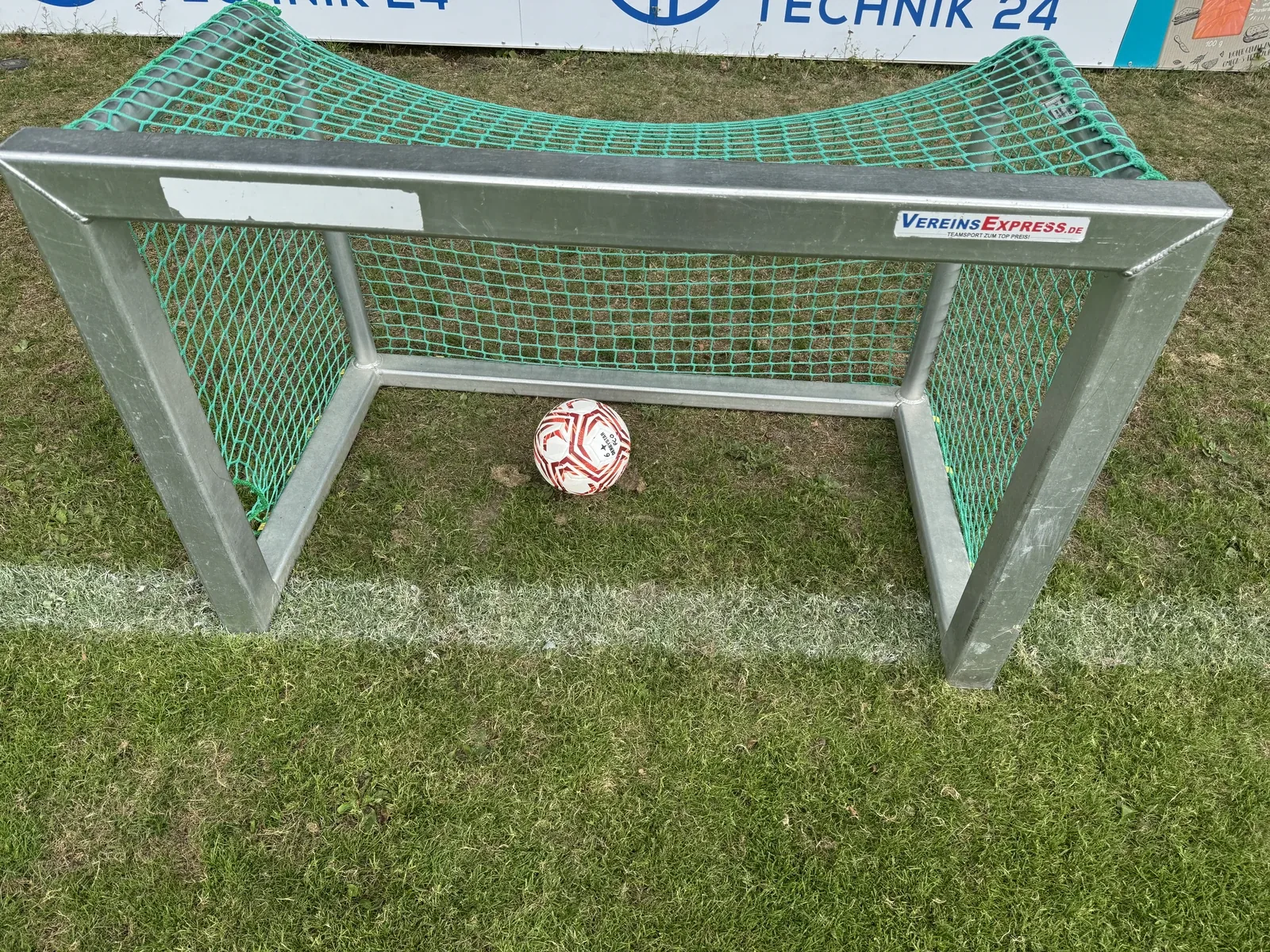 Ein verzinktes Minitor aus Aluminium-Vierkantprofil mit grünem Netz steht frontal auf dem Rasen, die Pfosten auf einer weißen Feldlinie. Im Tor liegt hinten an der Bodenstange ein rot-weißer Fußball der Größe 4 mit dem lesbaren Schriftzug Derbystar. Im Hintergrund eine Werbebande.